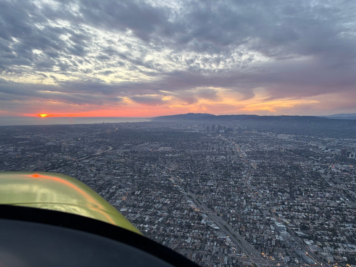 Yellow aircraft wing in the corner of the frame above Los Angeles streets at dusk, the sun setting over the ocean