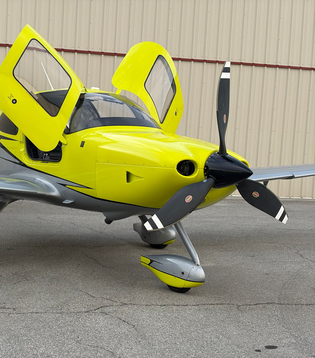 Front-quarter view of a yellow Cirrus SR22 with its doors open and propeller in front of a tan hangar