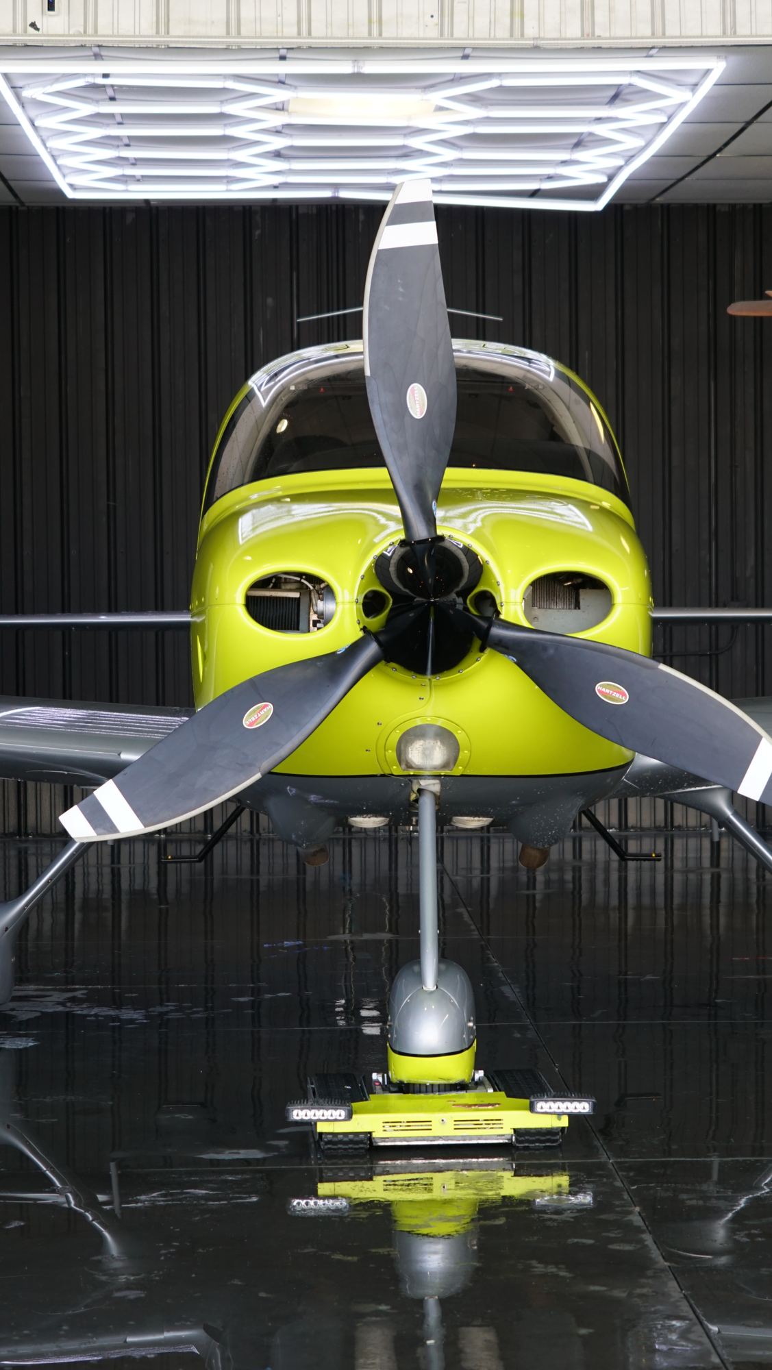 Yellow Cirrus SR22 nose-on inside a dark hangar, three-blade propeller and gear reflected in the polished floor