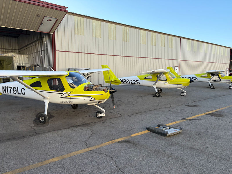 Three yellow Cessna 162 Skycatcher trainers in a row beside a hangar under a clear sky at Van Nuys Airport