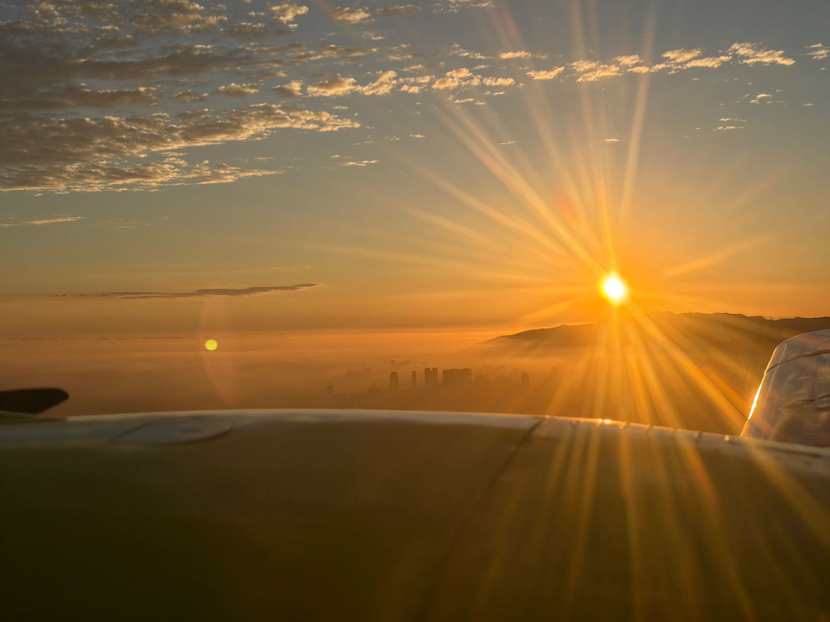 Low sun bursting over hills with the distant Downtown Los Angeles skyline in silhouette, seen over an aircraft nacelle