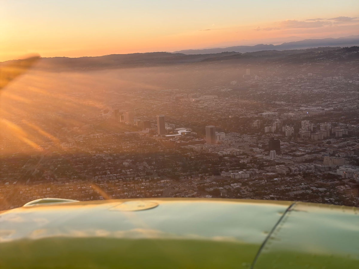 Sun flare streaming across hazy Los Angeles neighborhoods and hills, seen from the air over a yellow nacelle