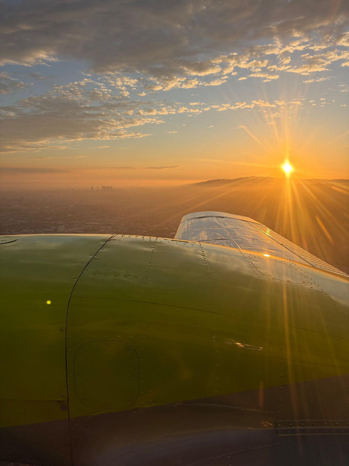 Sun setting over the hazy Los Angeles basin seen past the yellow engine nacelle of a Piper Seminole in flight