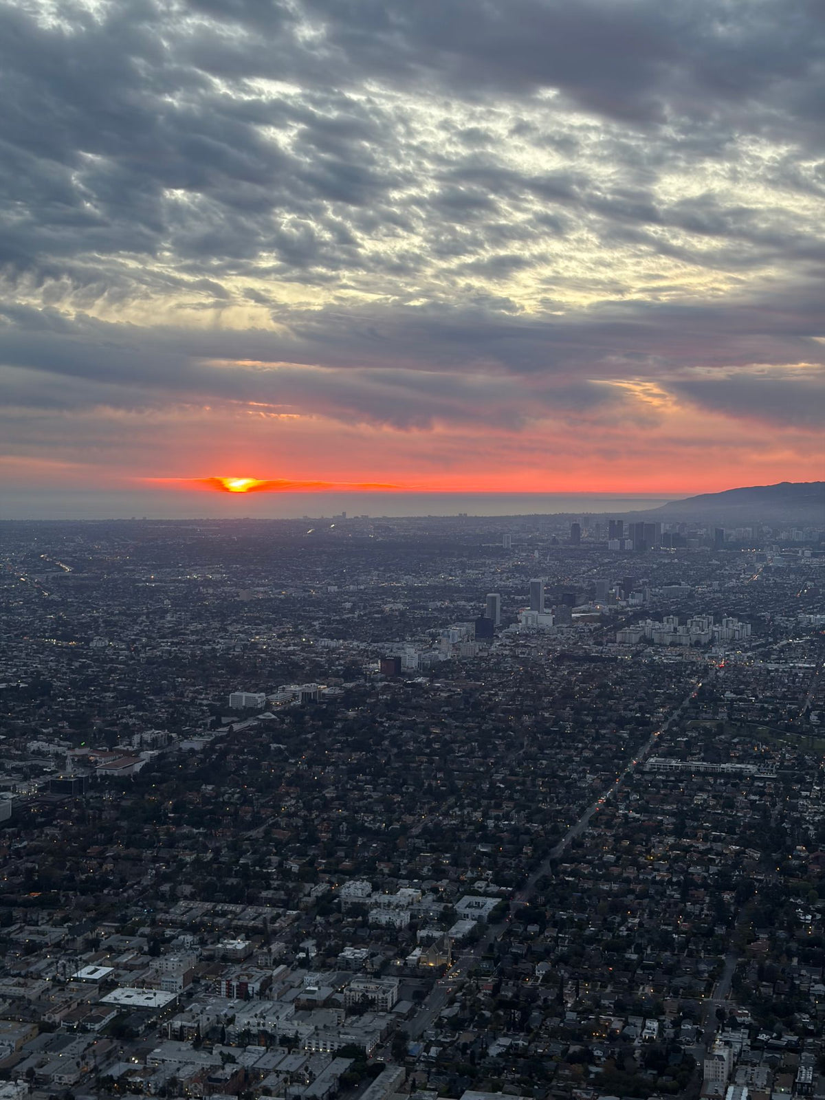 Aerial view at sunset across Los Angeles neighborhoods toward the ocean, with the sun on the horizon under a cloudy sky