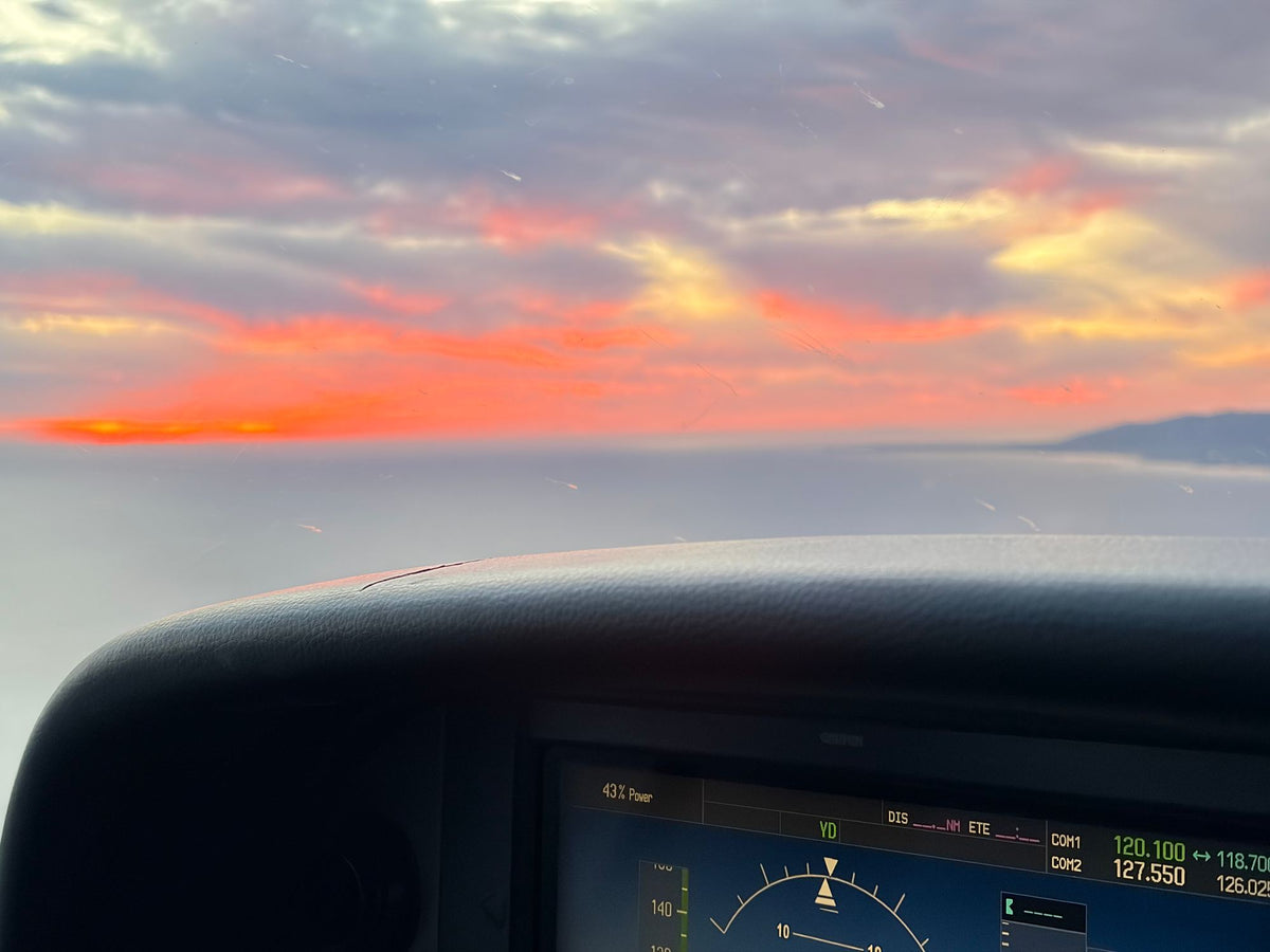 Glass primary flight display with attitude indicator and radio frequencies below a sunset sky over the ocean