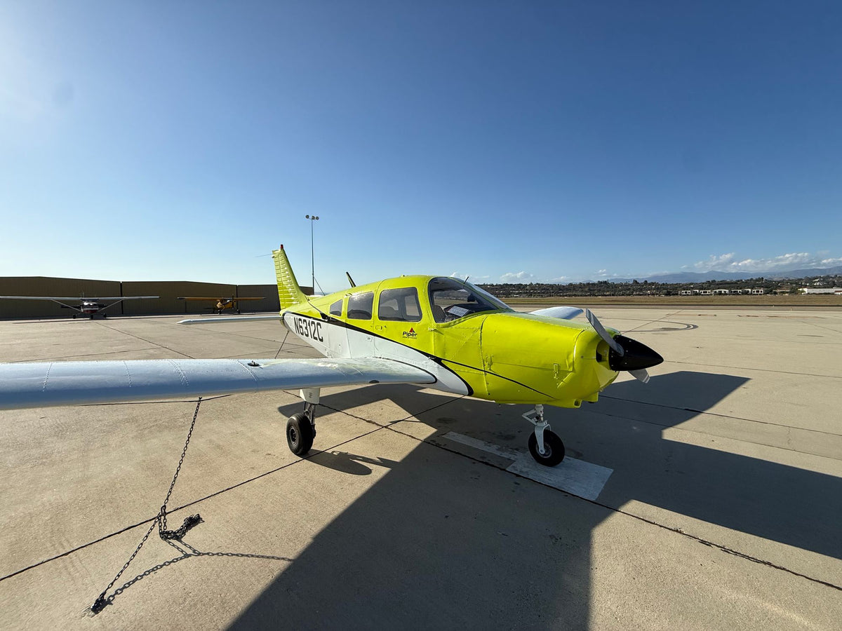 Yellow Piper Warrior N6312C seen from the right front quarter with its tie-down chain on a sunny ramp